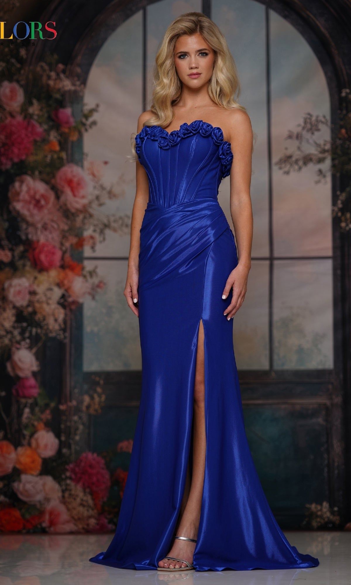 A woman poses before a floral backdrop in the Strapless Long Rosette Prom Gown (Colors Dress 3895) in Royal, featuring rosette details, a high slit, and an elegant strapless design.